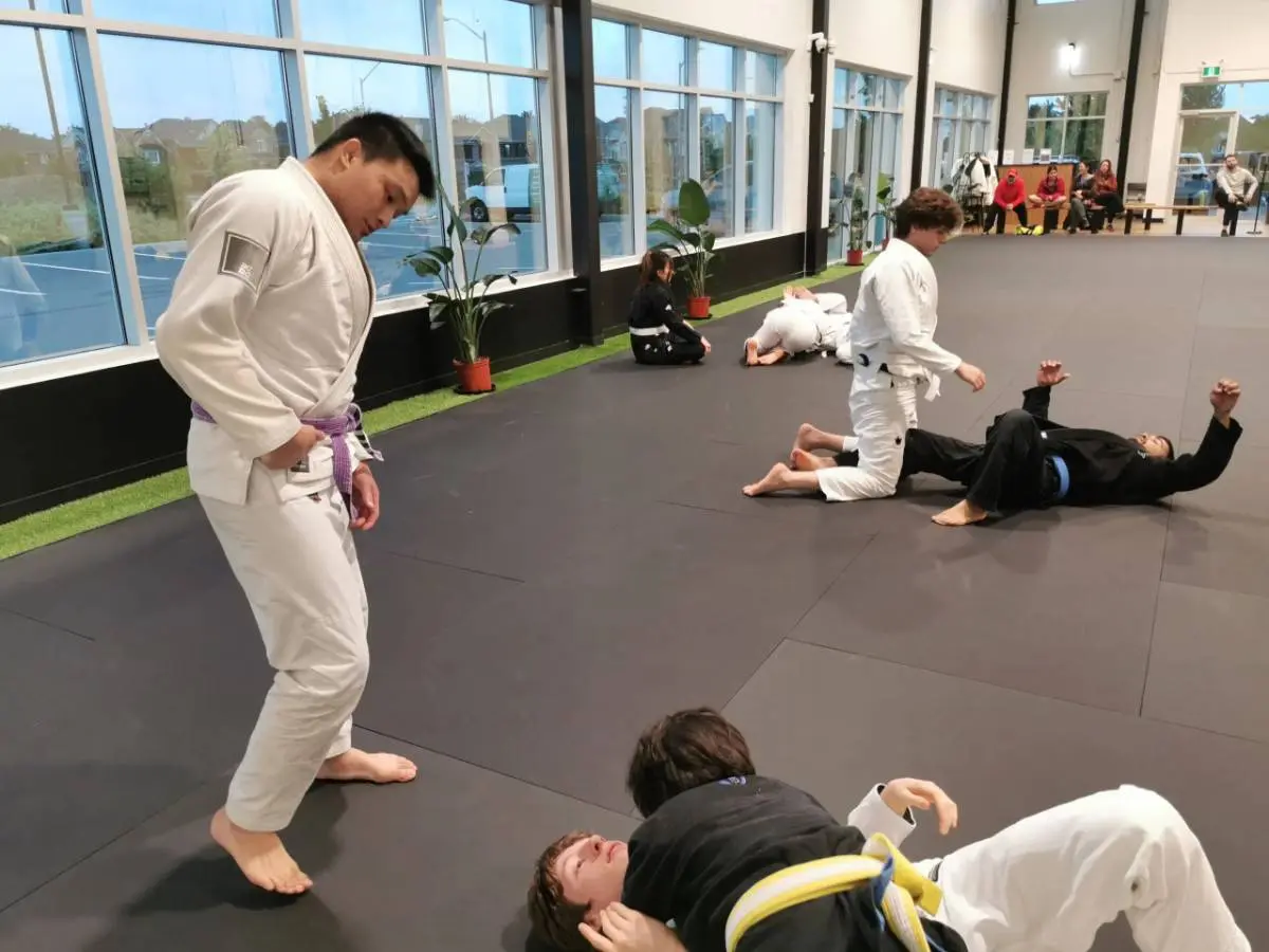 Multiple pairs of students in a large, modern martial arts facility practicing groundwork and sweeps during an adult judo class.