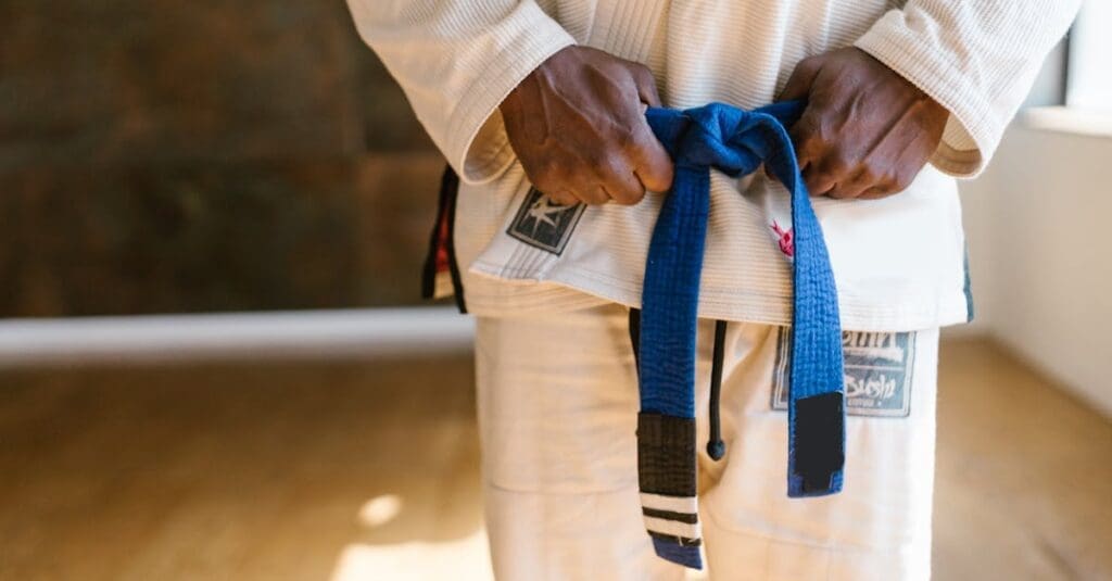 A close-up shot of a practitioner wearing a white gi tying a blue belt around their waist, a key milestone in learning Brazilian Jiu Jitsu Basics.