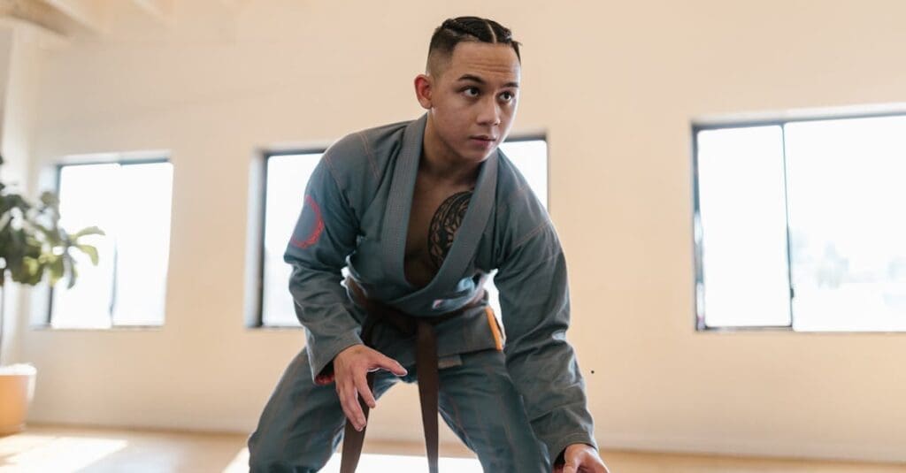 A focused martial artist in a grey gi and brown belt assumes a low, athletic stance on the mats while learning Brazilian Jiu Jitsu Basics.