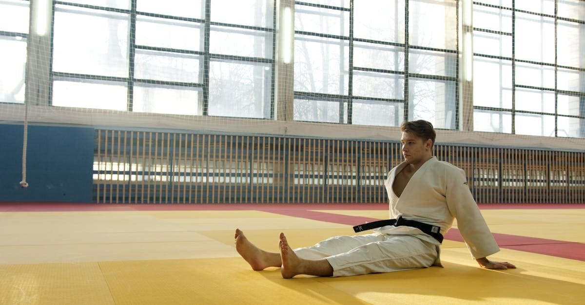 A male martial artist wearing a white judo gi and black belt sitting on a yellow and red tatami mat inside a gym with large windows.